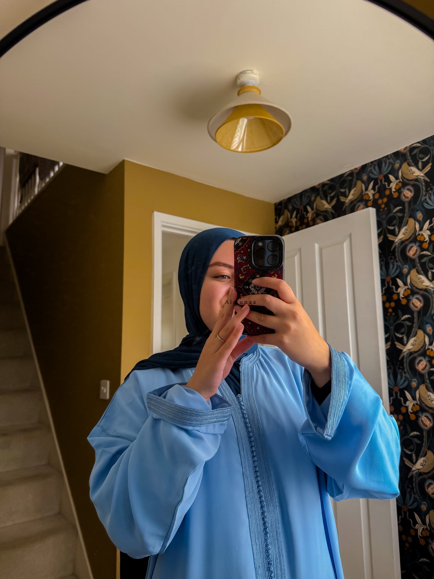 Person wearing a blue outfit taking a mirror selfie in a room with yellow walls and floral wallpaper.