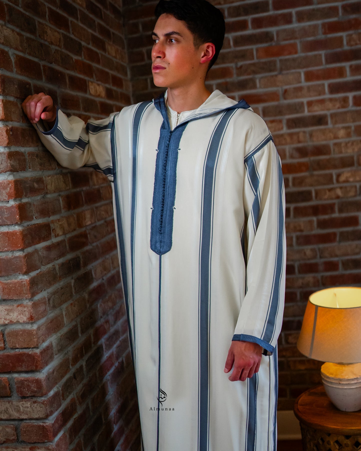 Man wearing a traditional outfit with blue accents against a brick wall.