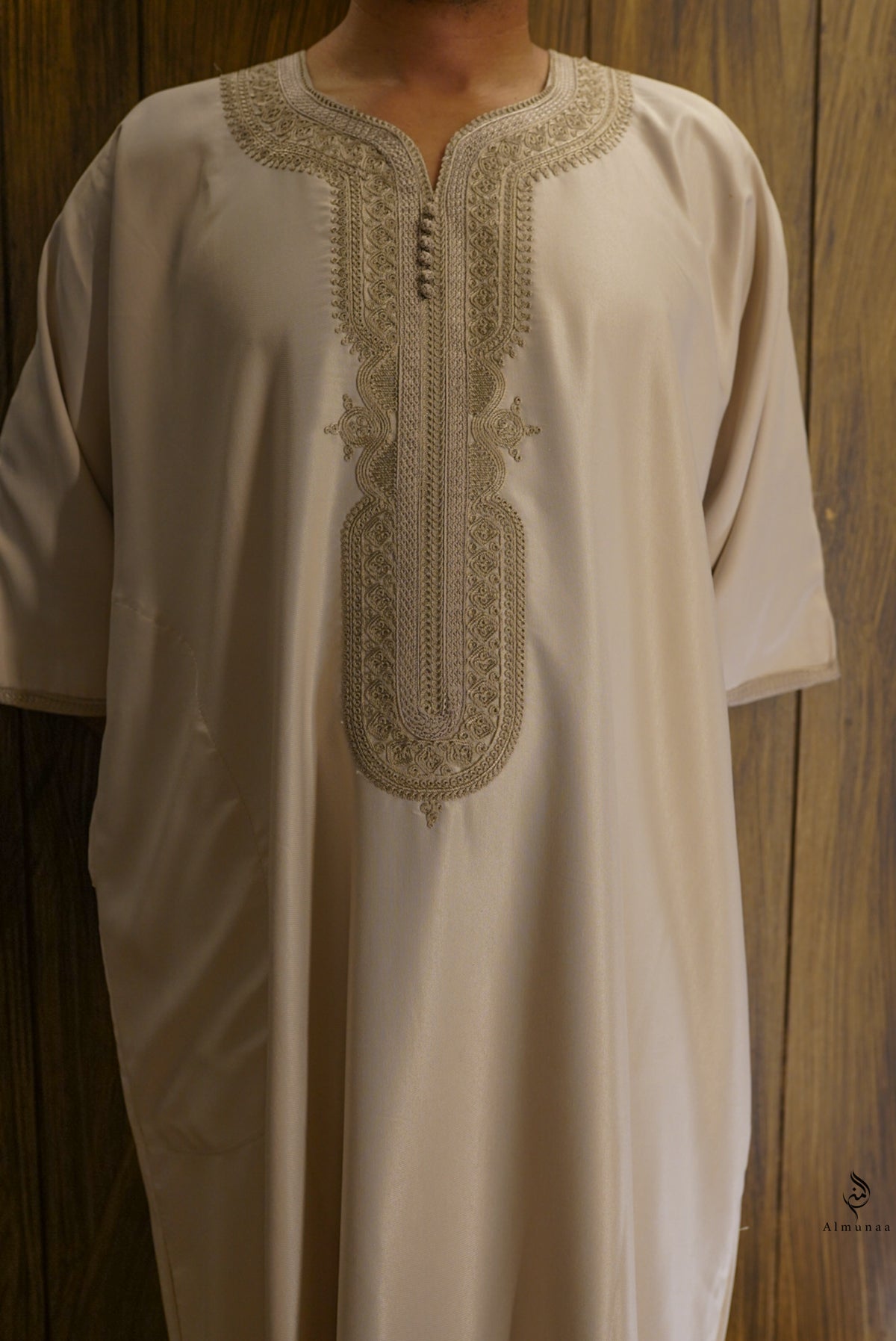 A cream-colored traditional long tunic with intricate beige embroidery around the split neckline and front placket, shown on a person against a dark wood panel background.