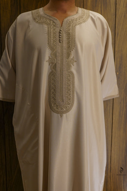 A cream-colored traditional long tunic with intricate beige embroidery around the split neckline and front placket, shown on a person against a dark wood panel background.
