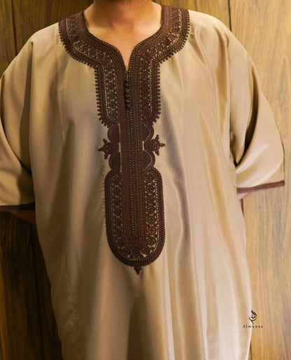 A beige short-sleeved tunic with intricate dark brown embroidery around the split neckline and center placket, shown on a model against a wooden background.