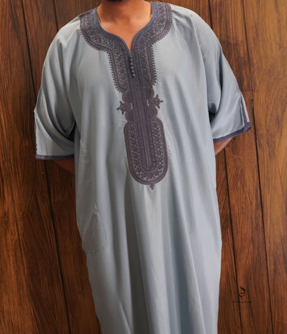 A person wearing a light gray traditional tunic with dark gray embroidered detailing around the split neckline and center chest, standing against a wooden plank background.