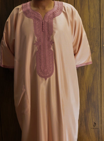 A peach-colored kaftan tunic with dusty rose embroidery on the neckline and sleeve cuffs, shown against a dark wood panel background.