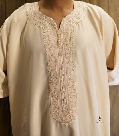 A close-up of a cream-colored tunic with intricate beige embroidery around the split neckline and front placket, worn by a model against a dark wood background.
