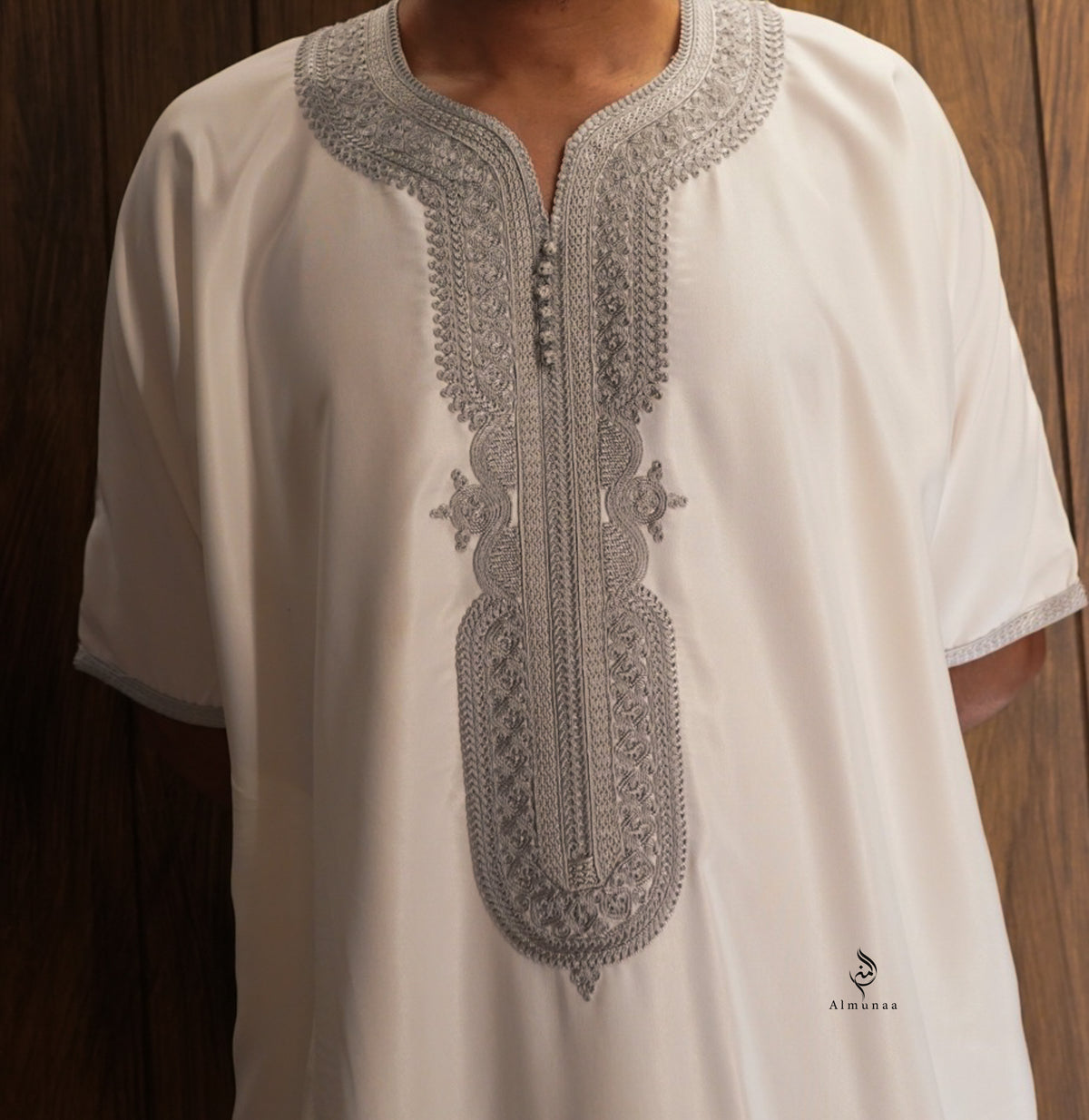 A white short-sleeve tunic with intricate silver-gray embroidery around the split neckline and down the center front, worn by a model against a dark wood background.