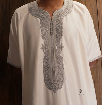 A white short-sleeve tunic with intricate silver-gray embroidery around the split neckline and down the center front, worn by a model against a dark wood background.