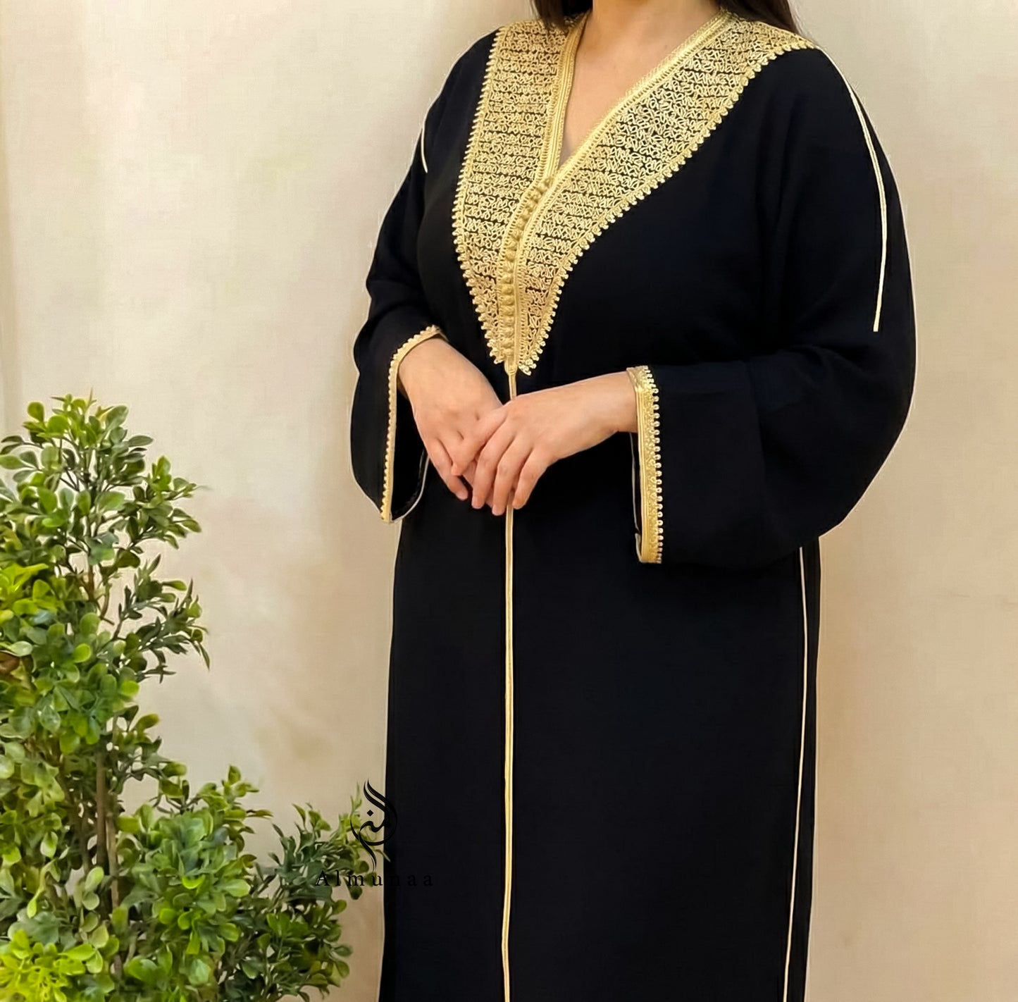 Person wearing a black dress with gold embroidery against a beige wall.
