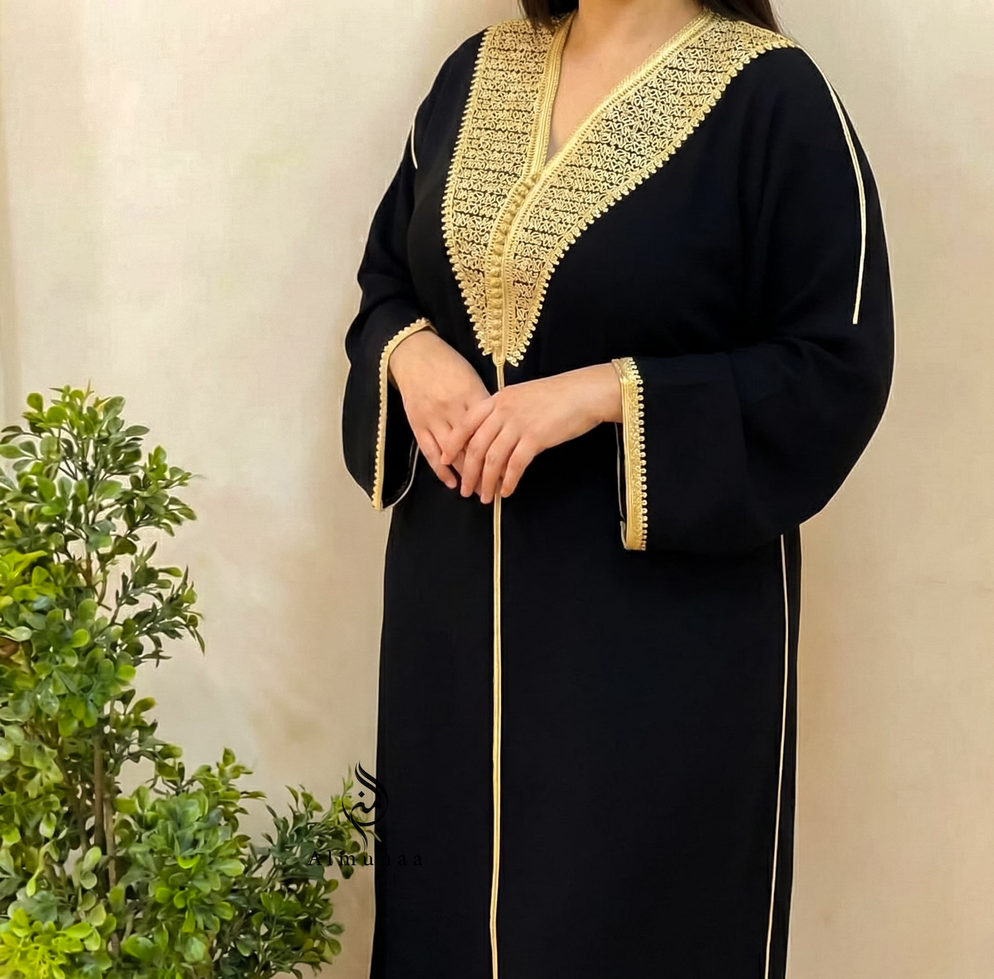 Person wearing a black dress with gold embroidery against a beige wall.